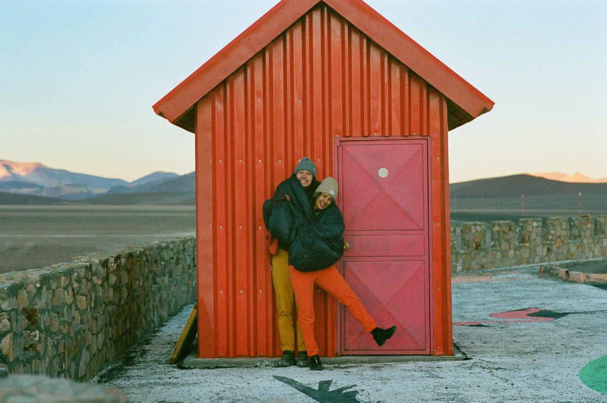 A color photo by Adam Wells of two models embracing in front of a small red building.
