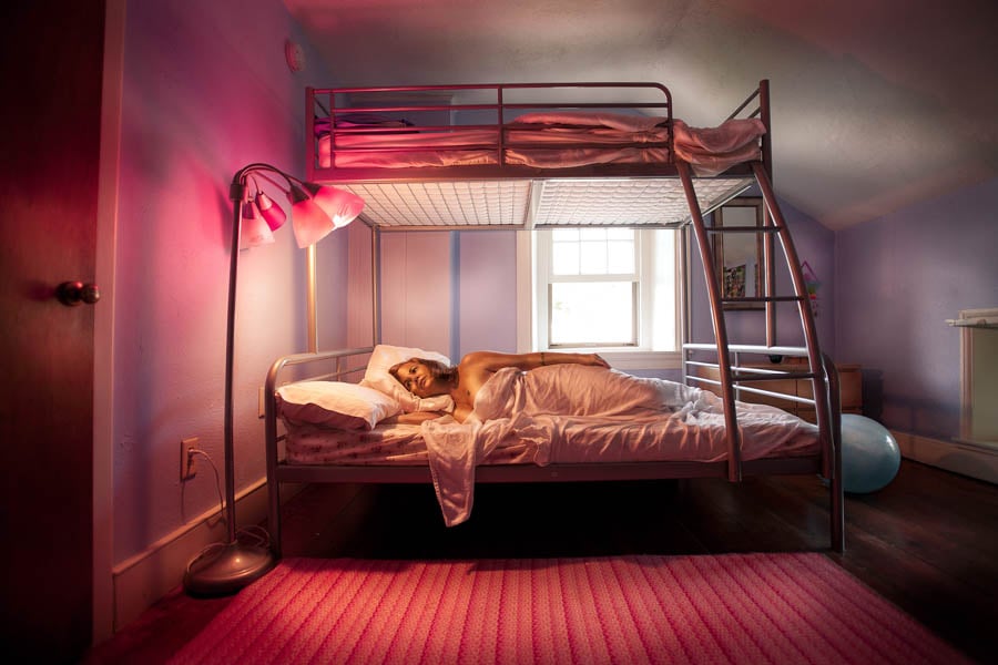 A woman lays naked in the bottom of a bunkbead, her room decorated in pink, from photographer Dave Moser's project "An American House Wife".