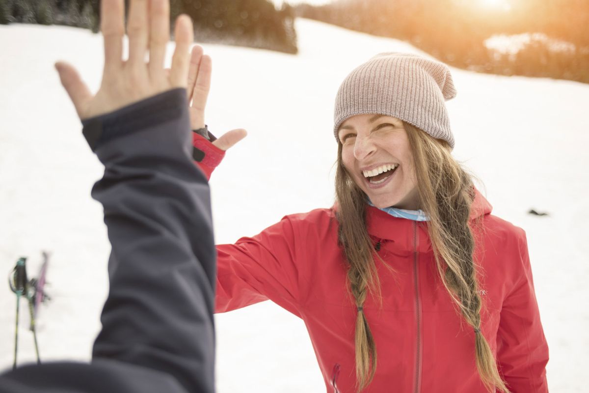 A woman with a beaming smile on her face engages in a spirited high-five with someone against the breathtaking backdrop of snow-covered mountains.