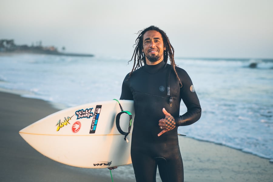 A smiling man holding his surfboard throws a shaka on the beach by photofrapher John Davis of Los Angeles, California