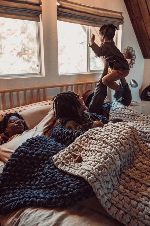 A young boy jumps in bed in his parents' as his sibling looks up and his other parent looks tired by photographer Michelle McSwain of Culver City, California 