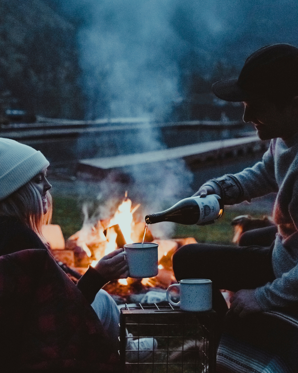 A man elegantly pours a cup of Kobrand wine for a woman, both dressed in cozy winter attire enjoying the warm ambiance of a crackling fire.