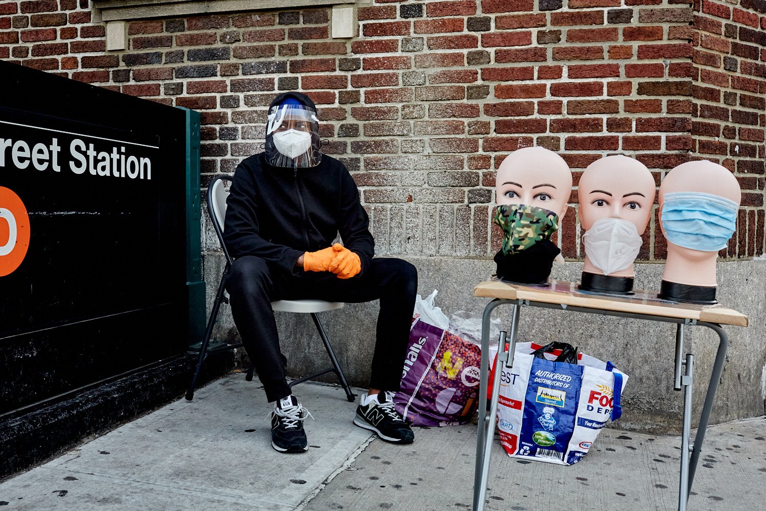 Man selling masks, sitting masked and faceshielded outside a subway station, by Stephen Speranza