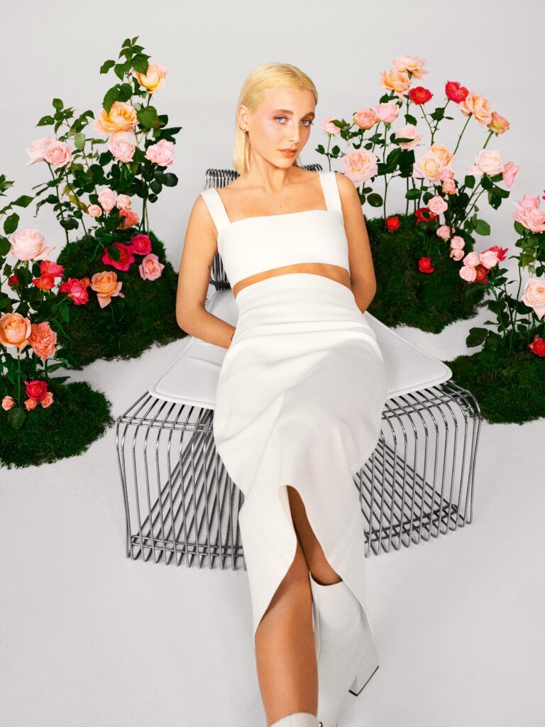 Dressed in a white dress and boots, YouTube star Emma Chamberlain poses on a modern chair surrounded by flowers.