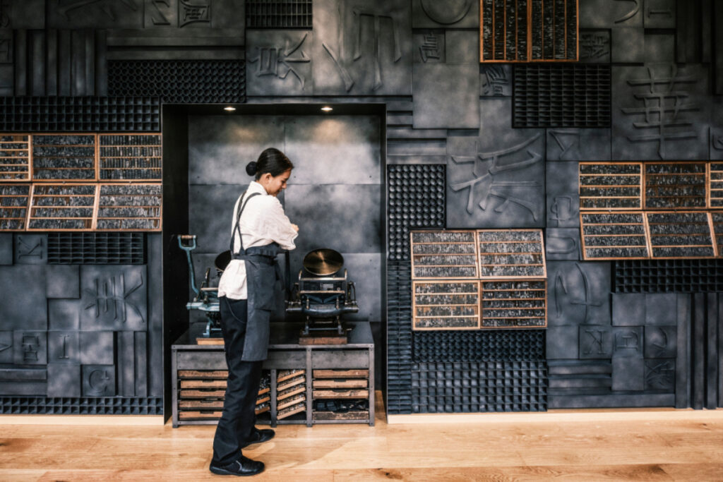 In a modern artisanal setting, a woman in a dark apron stands next to a machine, surrounded by shelves of type blocks, against a backdrop of an industrial wall embossed with Asian characters and geometric patterns.