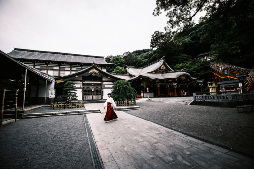 A graceful woman adorned in a white and red traditional Japanese ensemble strolls with poise, while the backdrop reveals the timeless charm of traditional Japanese architecture. 