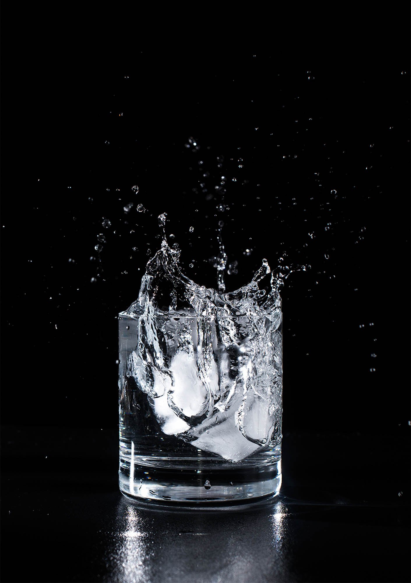 A color photo by Cayla Zahoran of ice splashing into a rocks glass full of water against a black background.