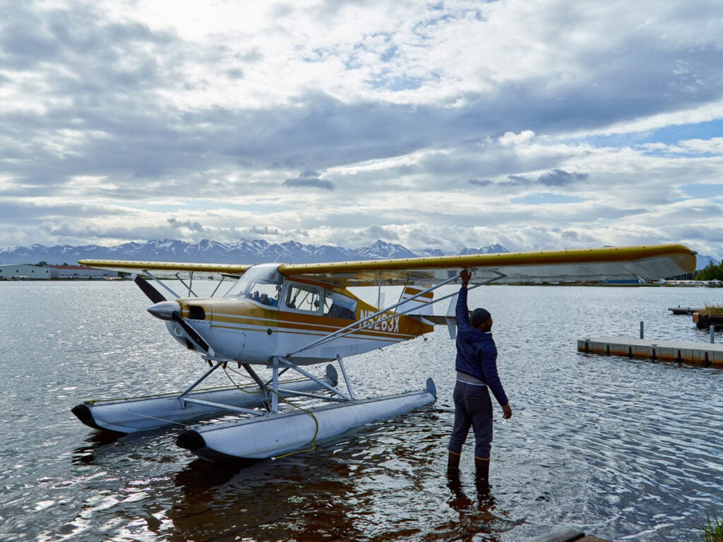A color photo of someone standing beside an amphibious airplane parked  on a lake by Clay Cook.