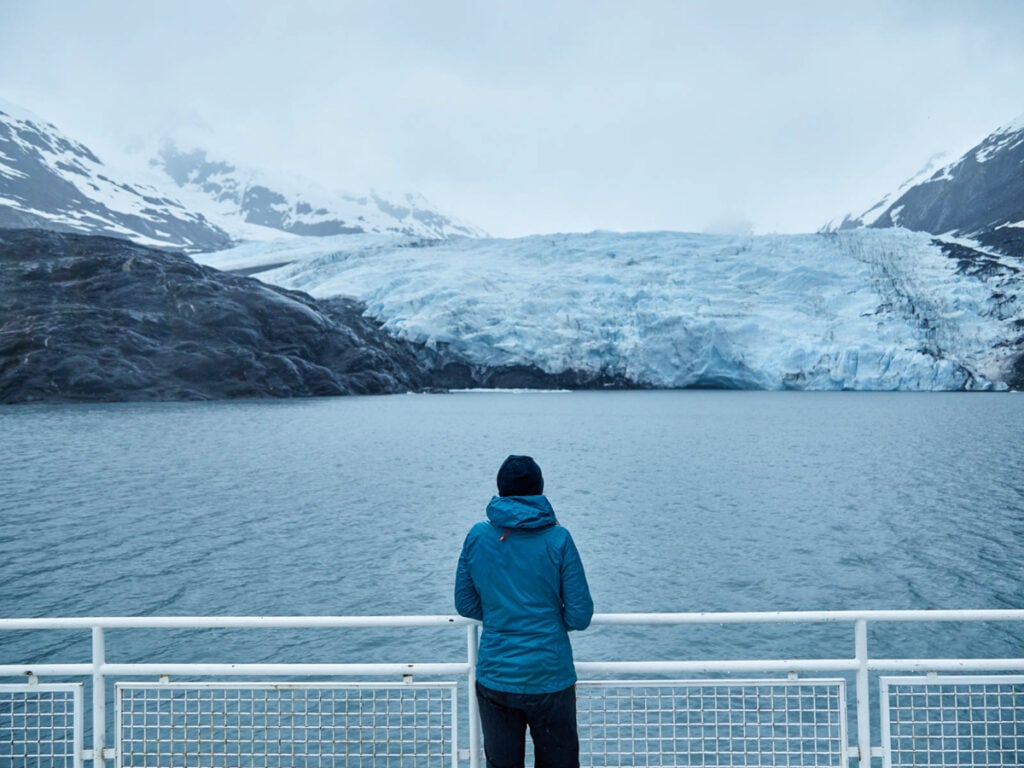 A color photo of someone taken from behind standing at a look out point across the water from an iceberg by Clay Cook.