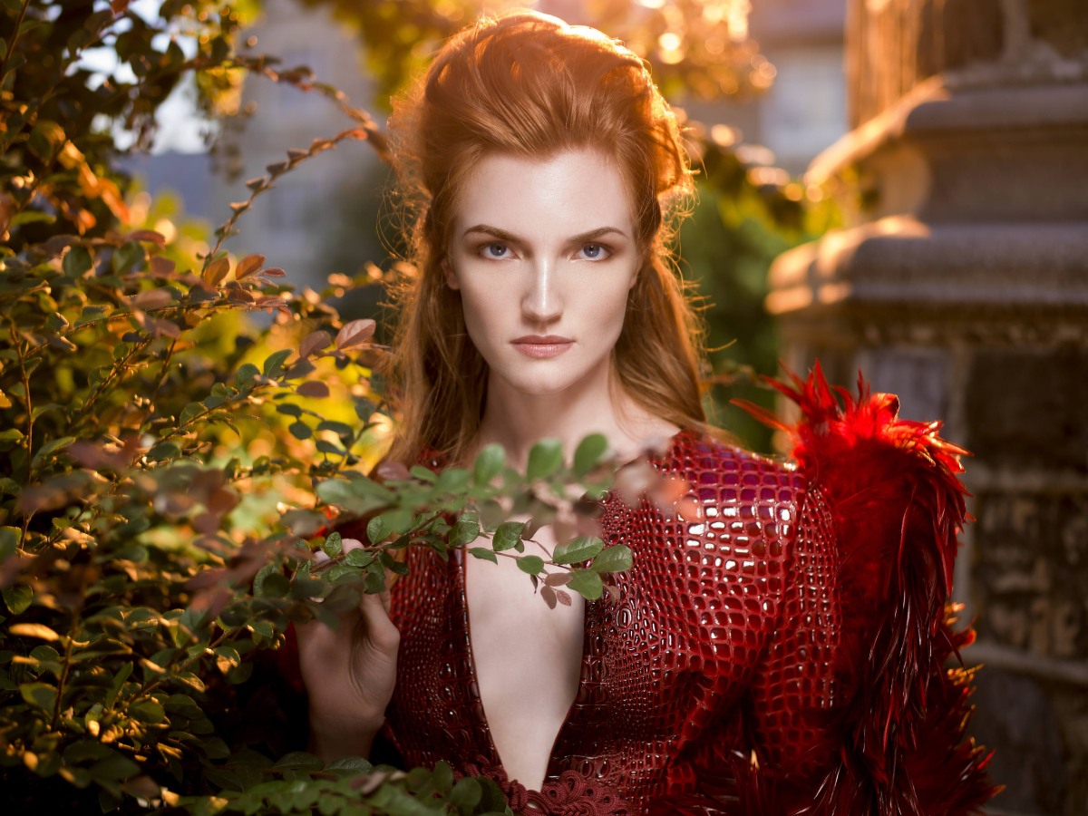 In a striking display of bold fashion, a woman confidently poses in a red leather alligator skin blazer adorned with vibrant red feathers. The natural surroundings enhance both her beauty and the audacious choice of attire, photo by Derek Blanks.