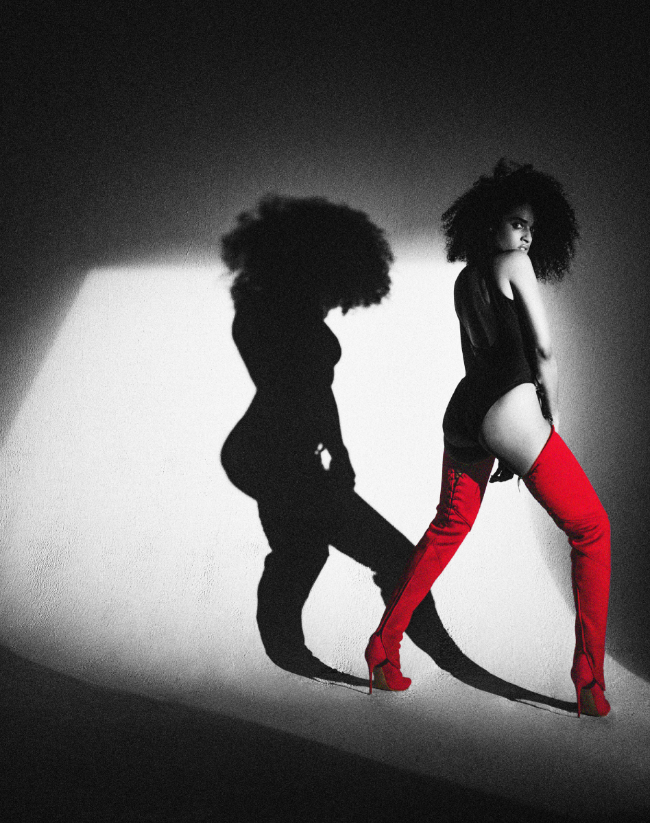 In a captivating black and white photograph, a woman dons a sleek black bodysuit paired with striking knee-high red boots that defy the monochrome palette. The entire image is bathed in grayscale, except for the vibrant pop of color emanating from the bold red boots, while skillfully placed lighting casts shadows that enhance the overall visual impact, photo by Atlanta photographer Derek White.