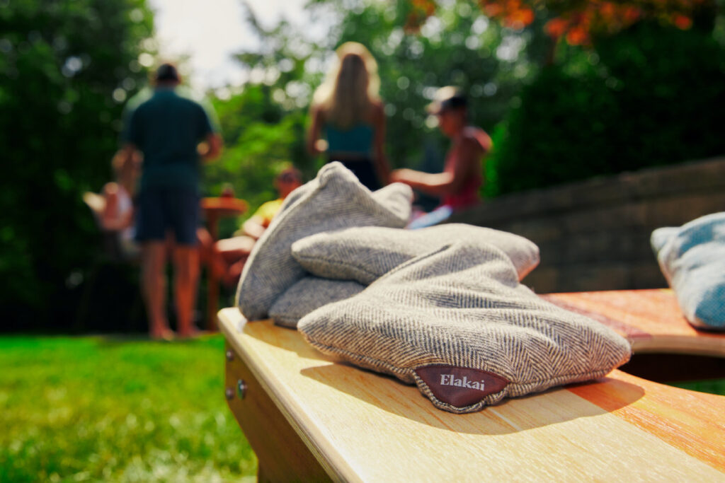 Devyn Glista's photograph of cornhole beanbags from the brand Elakai. The bags are resting on a cornhole board outdoors in the sun. Out of focus in the background is a group of people enjoying a get-together.
