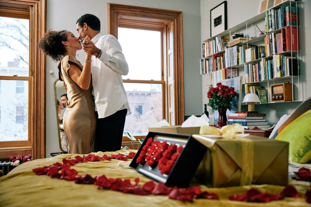 The image shows a loving couple dancing in their room. Red rose petals spell out "I love you" on the bed.