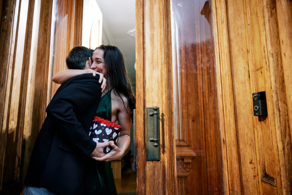 In the image, a woman stands at the doorway of her house, opening the door to a man. She warmly embraces him as he hands her a box bouquet of red roses, photo by Eli Meir Kaplan.