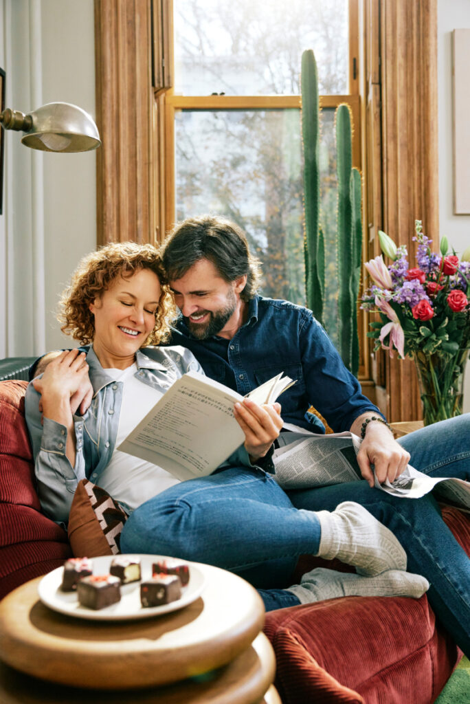In the image, a couple sits on the sofa, reading a newspaper together. They share a hug, smiling warmly at each other. In the background, a bouquet of beautiful flowers adds a touch of color to their cozy moment, photo by Eli Meir Kaplan.