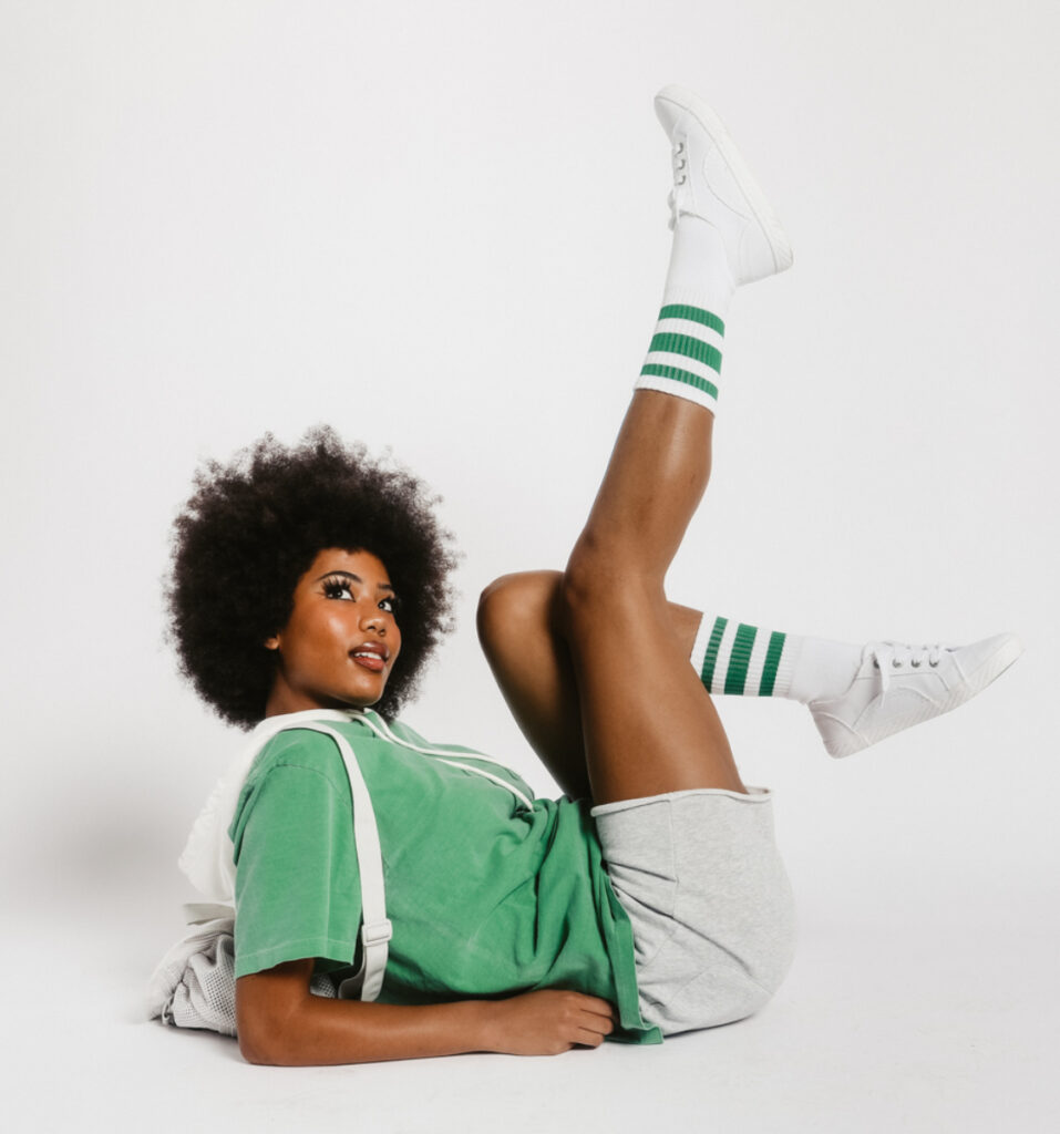 A photo by Erica Joan taken in a studio of a Black woman wearing a shortsleeved kelly green zip-up hoody, gray shorts, and white sneakers with socks pulled halfway up her shins. Her hair is styled in an Afro, and she poses on the ground, leaning back and propped up on her elbows, kicking her feet in the air playfully.