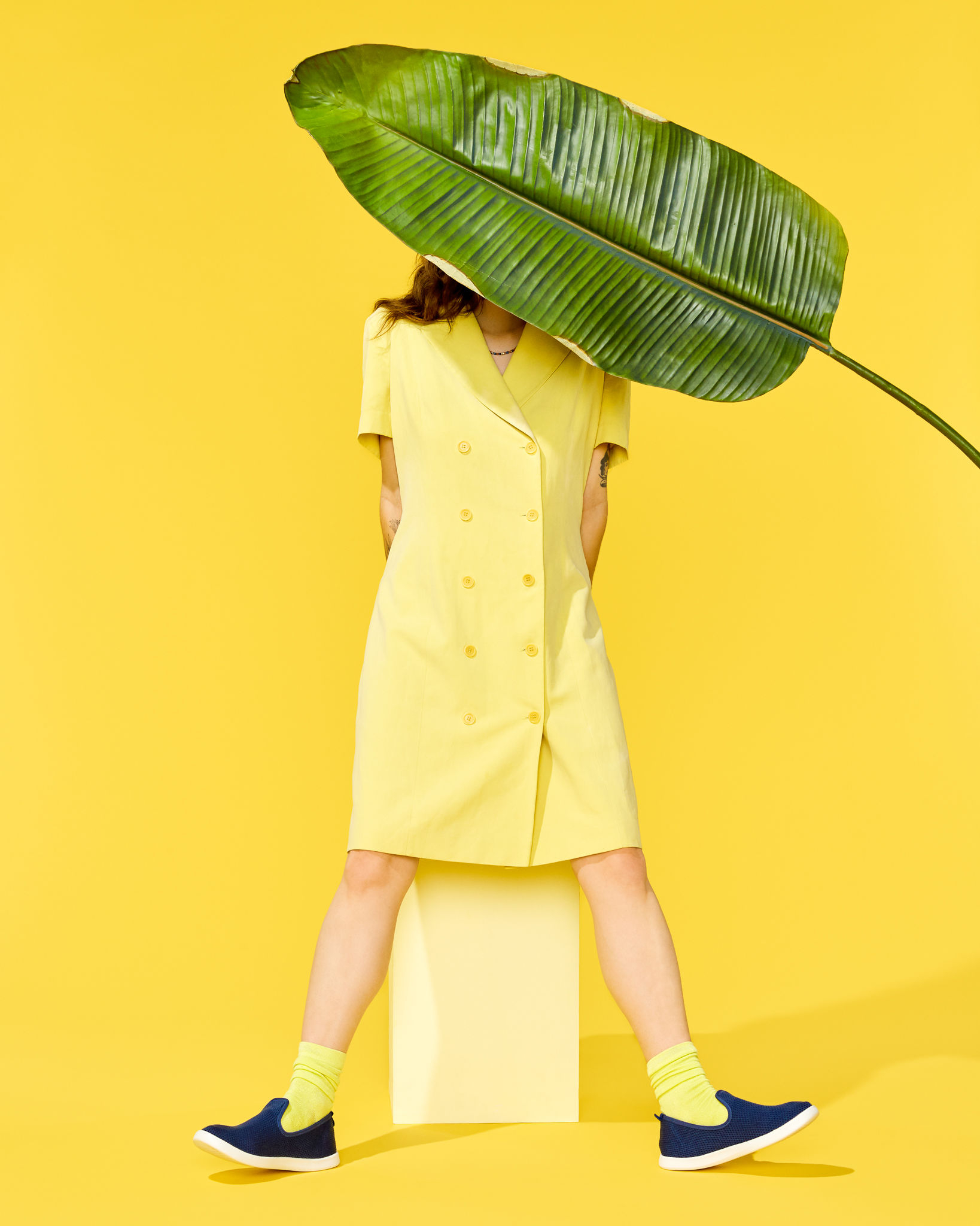 A woman elegantly presents a vibrant yellow dress, her head playfully concealed behind a large, lush green leaf. The background harmoniously echoes the radiant yellow tones, creating a captivating and visually cohesive composition, photo by TROPICO PHOTO.