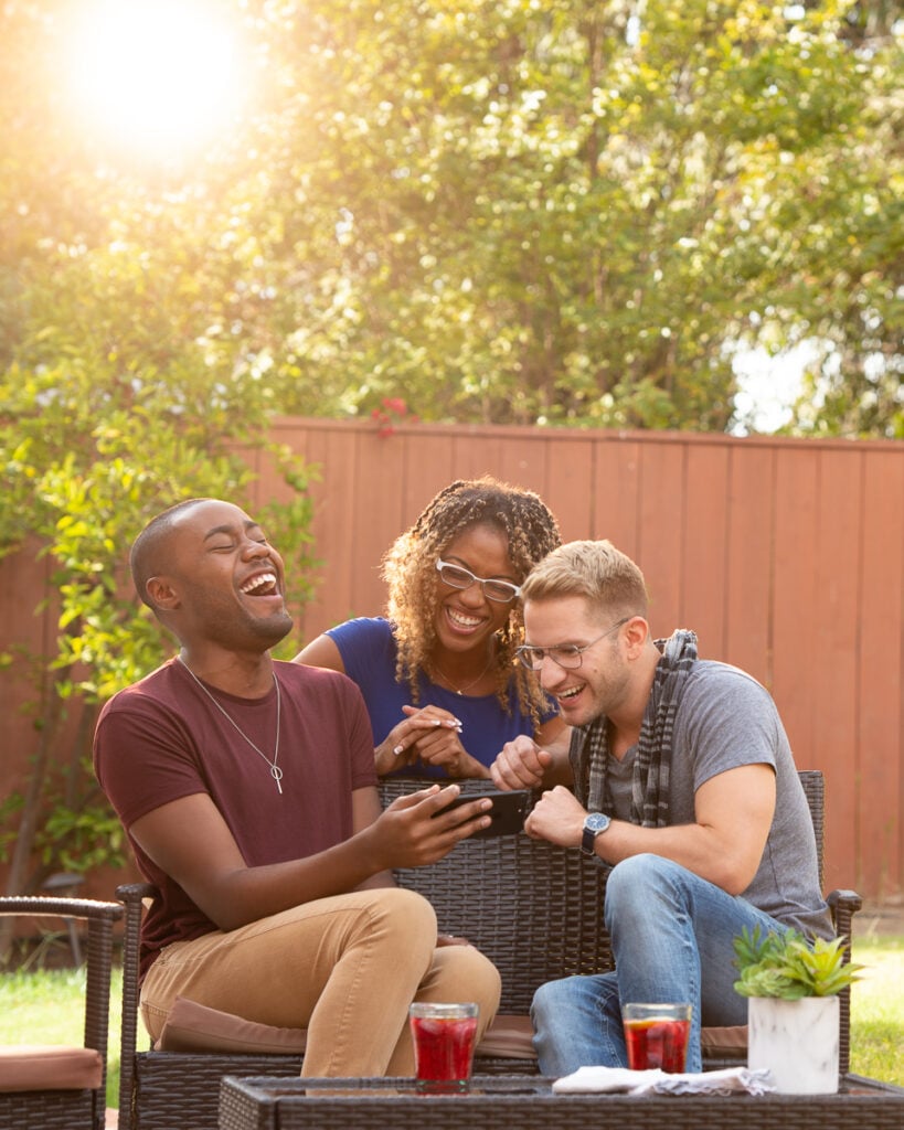 Three milennials laugh while watching a video on a phone in a sun soaked back yard, photographer by Frank Rogozienski.