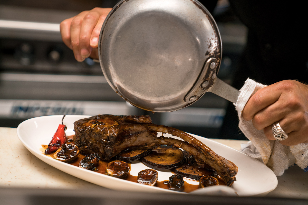 A skilled chef is artfully preparing a mouthwatering lamb cutlet, accompanied by succulent aubergine slices and perfectly cooked cherry tomatoes. The entire ensemble is skillfully glazed with a tantalizing sauce, creating a glossy and flavorful finish, photo by Gary Payne.