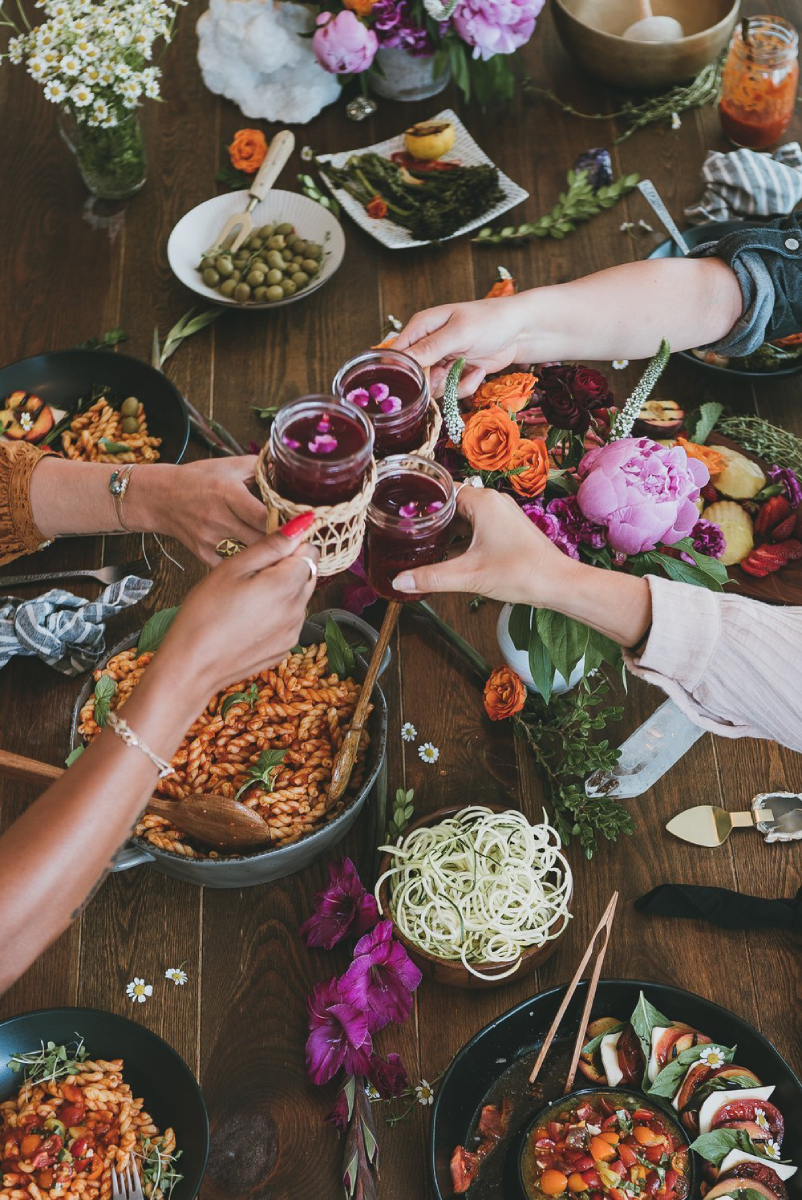 Image shows people toasting with a delightful pink drink. The table is adorned with a tempting feast, featuring a delectable tomato pasta dish and a fresh, inviting salad. The scene is not only a celebration of flavors but also a visual treat, with vibrant flowers elegantly enhancing the table's ambiance, photo by Hannah Bernabe.