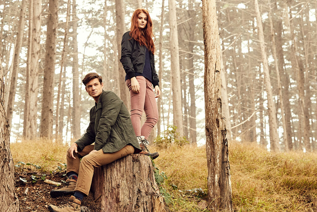 James Bueti's photograph of two models posed in a forrest in bright daytime. The male model sits on a stump and the female model stands next to him on the same stump. They both wear outdoorsy boots.