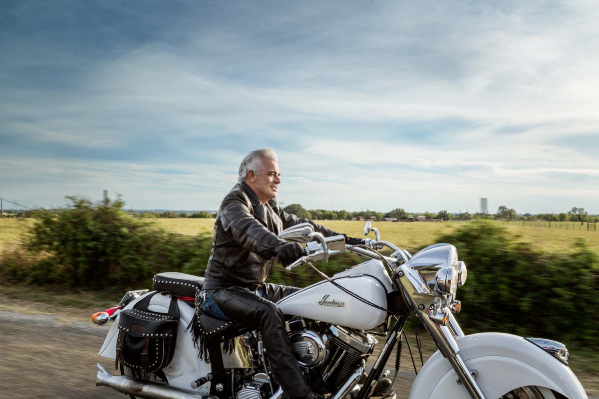 A color photograph by Jeff Wilson of a grey-haired man riding a motorcycle in the countryside without a helmet.