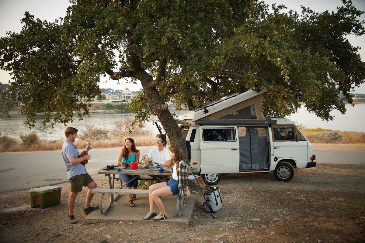 A color photograph by Jody Horton of friends sitting at a picnic while one of them plays a ukulele alongside a lake with their VW van alongside them.   