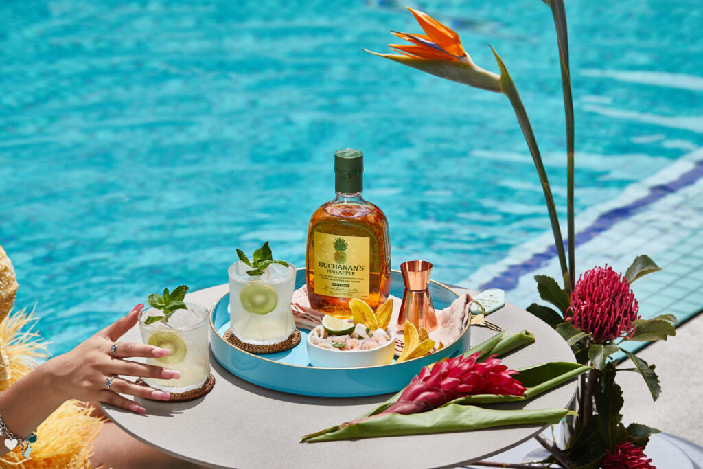 A bottle of Buchanan's Pineapple Whisky is elegantly displayed on a tray with a shot dozer and snacks, accompanied by two vibrant cocktails, sitting on a table near a pool.