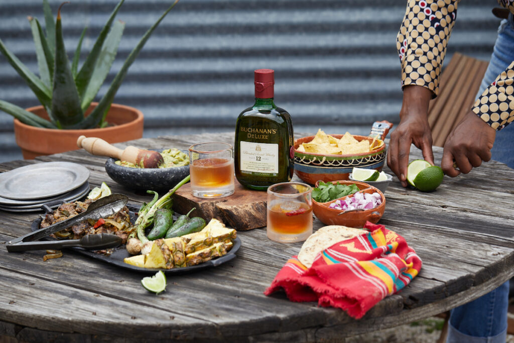 A bottle of Buchanan's Deluxe Whisky takes center stage on a rustic wooden table, its amber contents poured into two chilled glasses, as a man skillfully prepares a Mexican meal featuring an array of vibrant ingredients such as tortilla chips, onion, parsley, lime, guacamole, and roast veggies. Image by Jody Horton.
