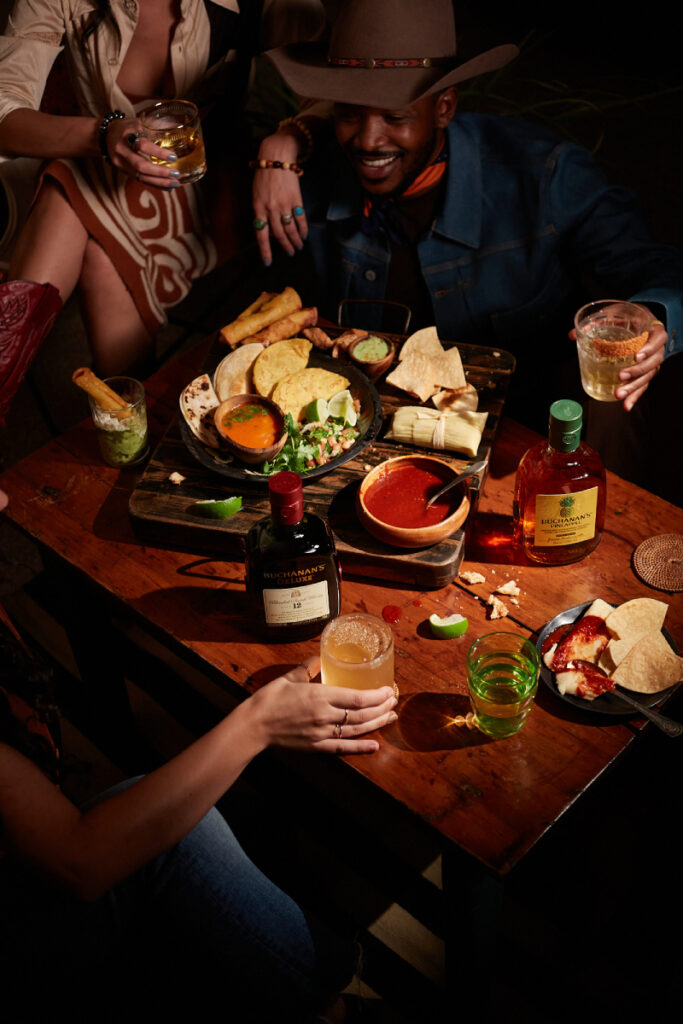 A man wearing a cowboy hat and women adorned in cowboy boots gather around a table, relishing a delicious meal accompanied by glasses of Buchanan's Pineapple and Deluxe Whiskey, enjoyed both straight and mixed into refreshing cocktails.  Photo by Jody Horton.