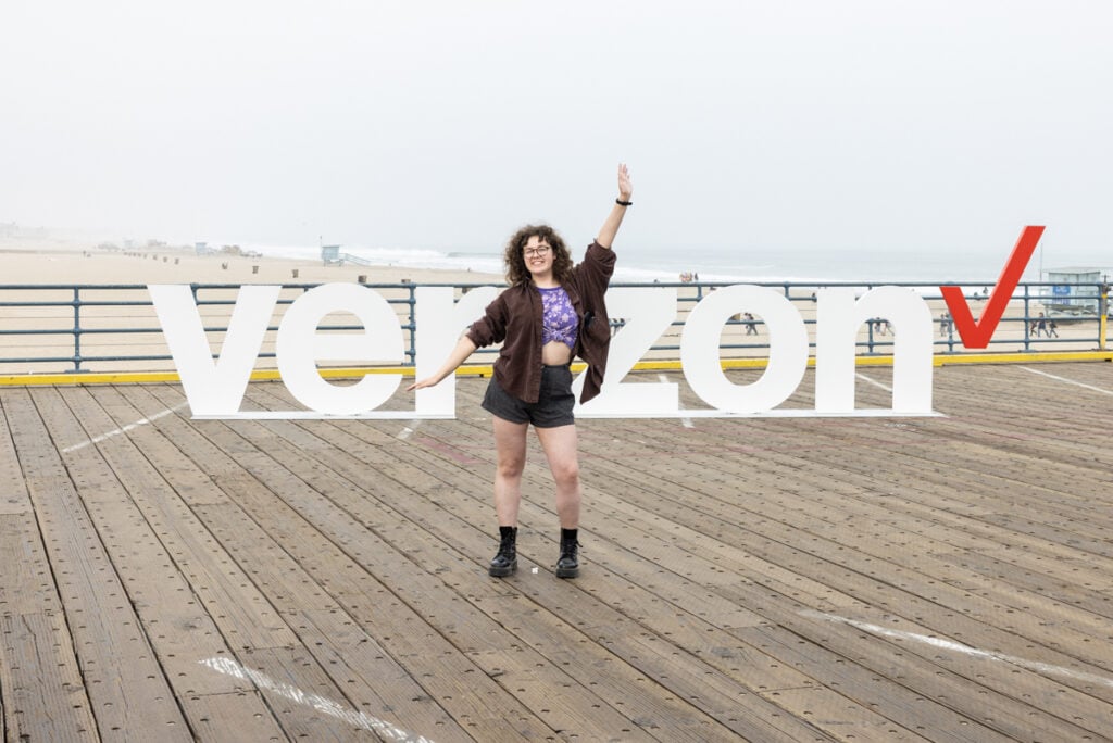 A photo by John David of a woman posing in front of the Verizon letters on the Santa Monica pier.