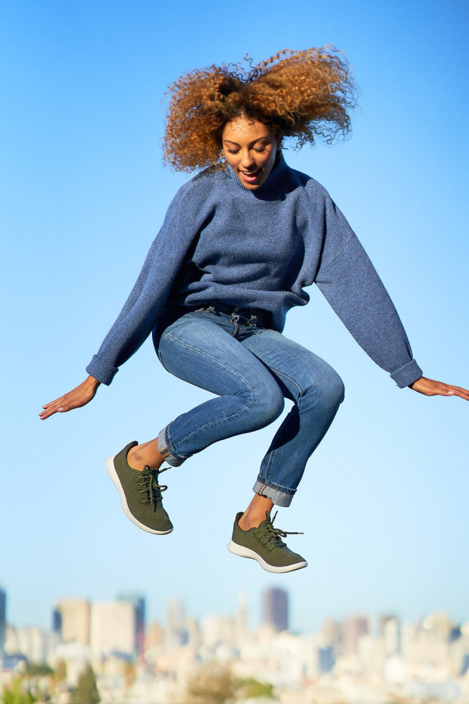 A color photo by Kris Cheng depicting a woman wearing blue jeans and a blue sweater jumping in the air against a blue sky and city skyline.