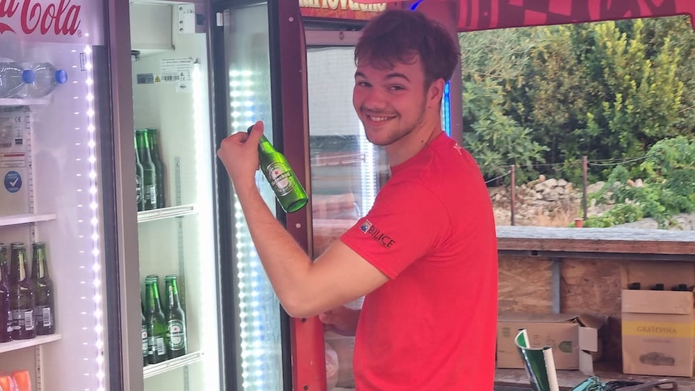 Portrait of Luka smiling in front of beer cooler, with bottle in hand.