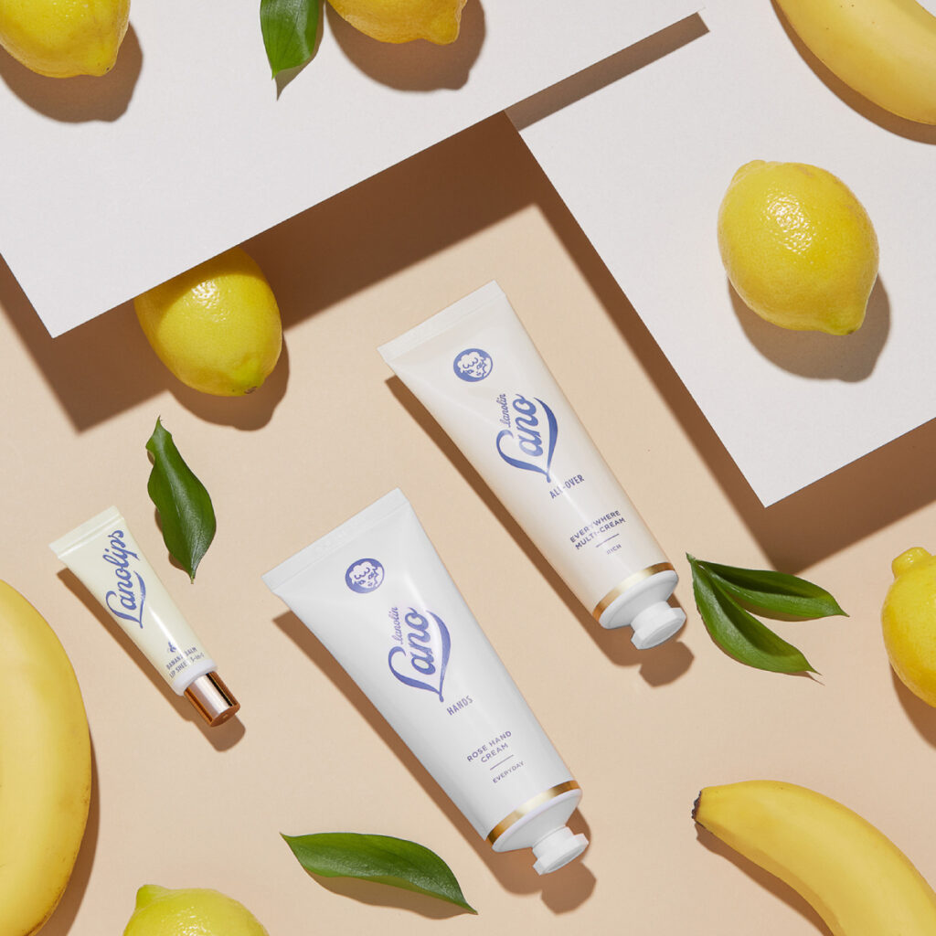 Cincinnati product photographer Marlene Rounds' image of three products from the brand Lanolips in tubes in various sizes. They are set against a light peach surface and are interspersed with lemons, bananas, and green leaves.