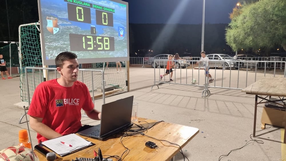 Photo of Mate sitting at announcer's table with computer and notepad watching game.