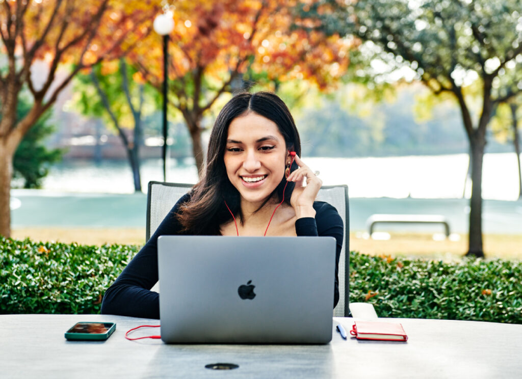 A girl sits on a terrace, wearing earphones and working on her laptop.