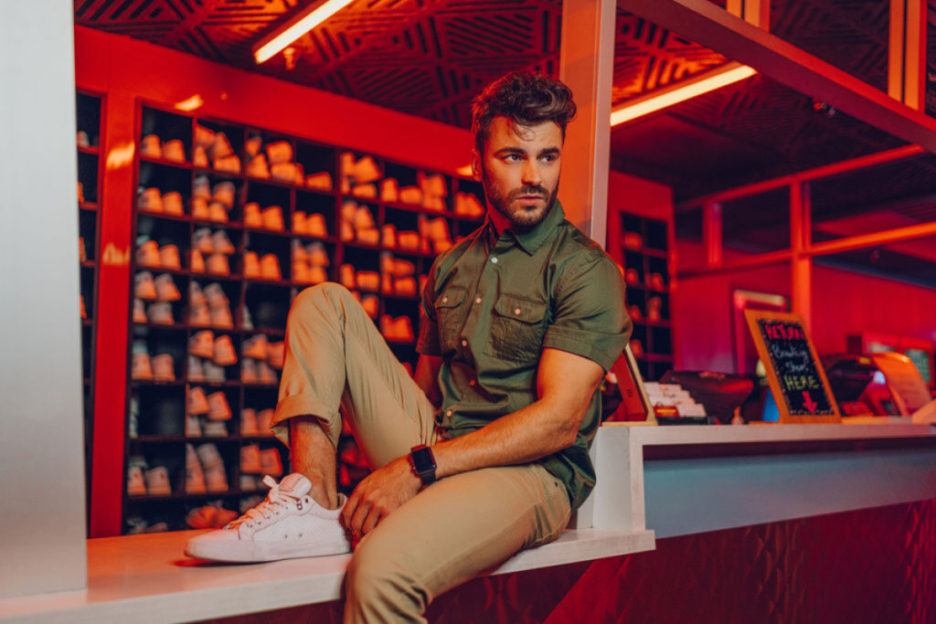 San Diego-based photographer Oveth Martinez's photo of a male model with facial hair wearing a sheeny moss-green short-sleeved cargo shirt, tan khaki pants, and white sneakers. He appears to be sitting on the counter of the shoe rental area at a bowling alley, and the room is lit with orange light.