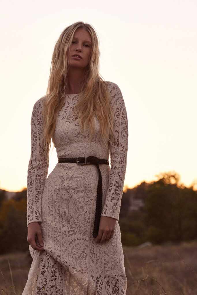 Fashion photographer Paula Watts' image taken outdoors at dusk of a model with long blonde hair wearing a white long-sleeved lace dress with a dark belt around her waist.
