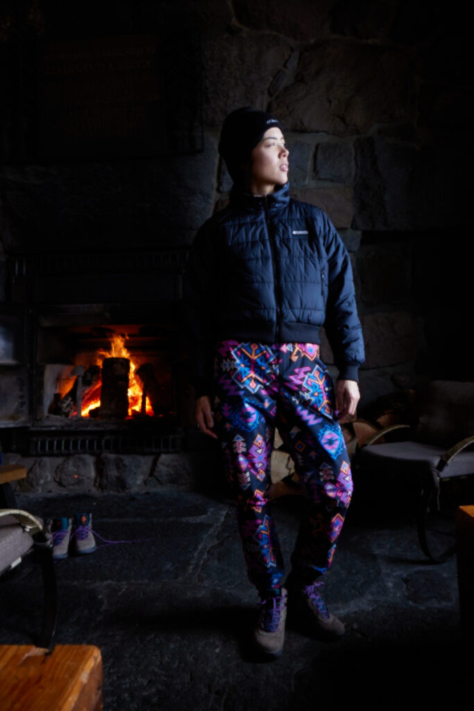 Dressed in the cozy attire of Columbia Sportswear's heritage collection, a woman strikes a pose in front of the crackling fireplace inside the ski lodge.