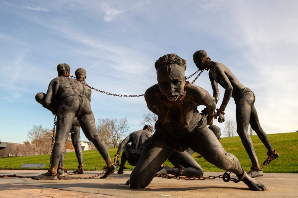 Image showing sculptures of black slaves adorned with chains around their necks, hands, and feet, their faces etched with palpable anguish and suffering, photo by Robert Rausch.