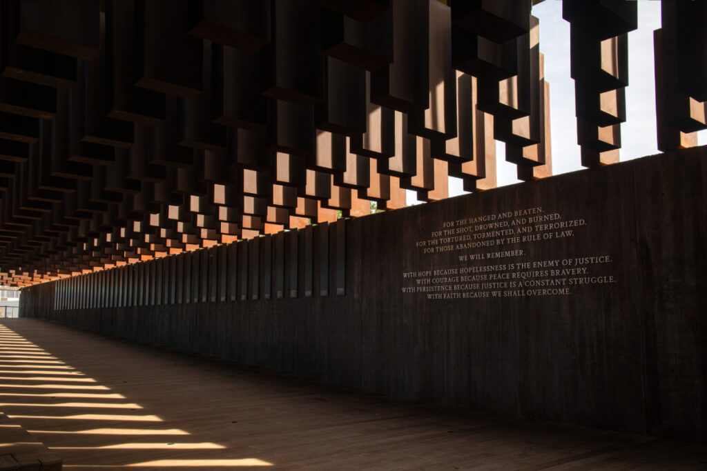 The image showcases the National Memorial for Peace and Justice featuring more than 800 corten steel monuments engraved with the names of the tortured. The Memorial features a poignant inscription: 'For the hanged and beaten, for the shot, drowned and burned. For the tortured, tormented, and terrorized. For those abandoned by the rule of law. We will remember. With hope because hopelessness is the enemy of justice. With courage because peace requires bravery. With persistence because justice is a constant struggle. With faith because we shall overcome.'