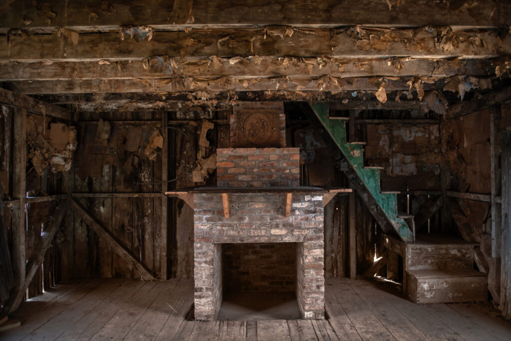 Image showing an inside of one of the wooden houses where slaves used to live. In the middle of  the hut is a fireplace and beside it a green staircase.