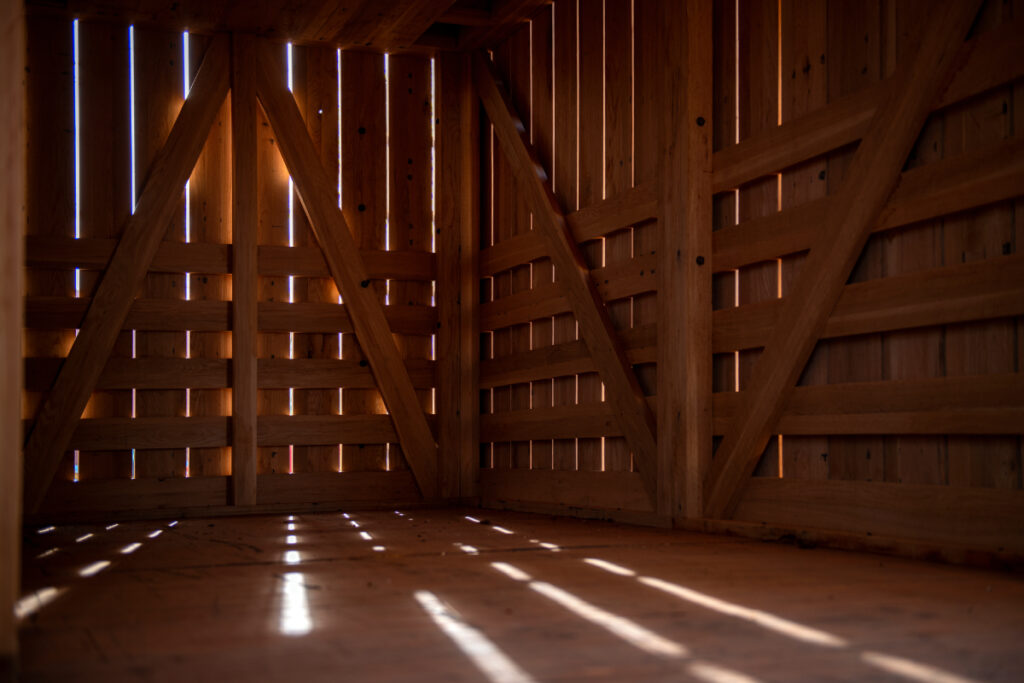 Image showing the inside of a wooden hut, the sunlight peeking through the cracks between the wooden boards and casting intricate patterns on the floor, photo by Robert Rausch.
