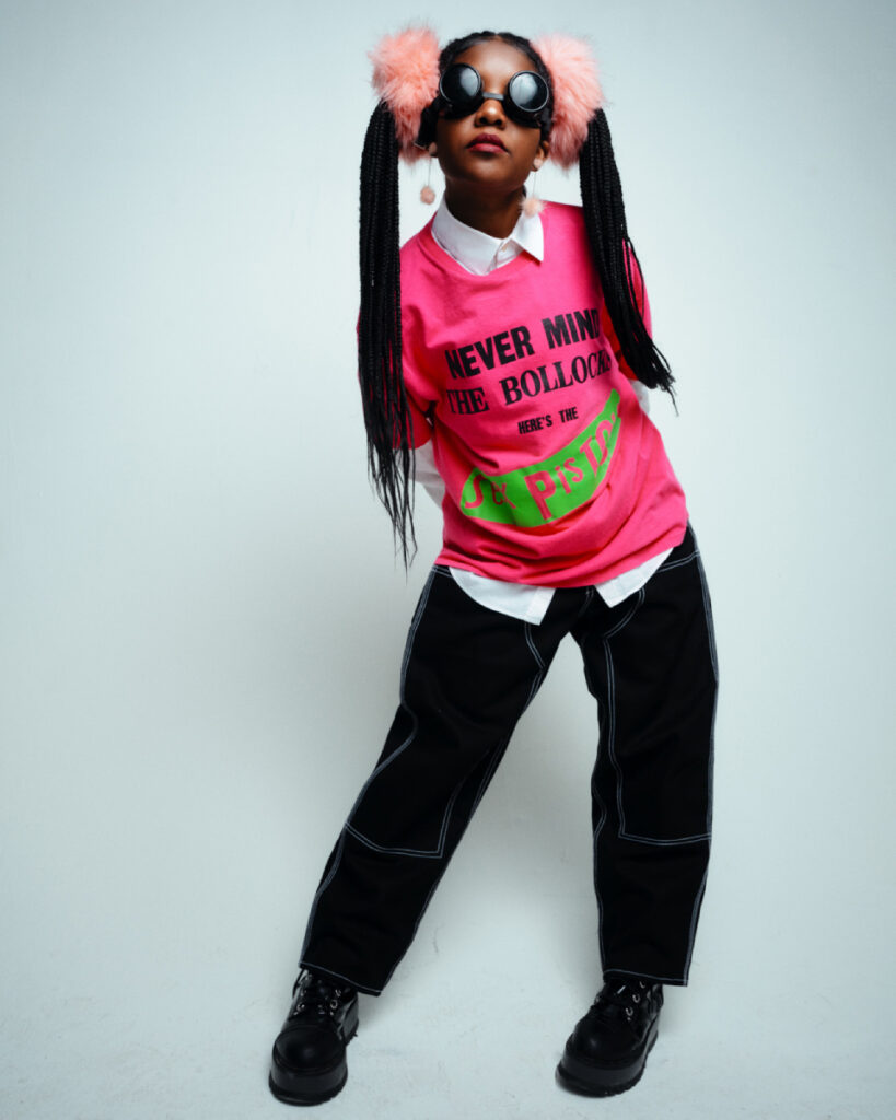 Ryan Allan's photo of a woman posing on a white studio set wearing a hot pink Sex Pistols t-shirt over a white long-sleeved button-down shirt. Her style is edgy. Her hair is styled in box braids that are arranged into two pigtails with furry light pink scrunchies, and she wears a pair of black round sunglasses that are reminiscent of swim goggles.