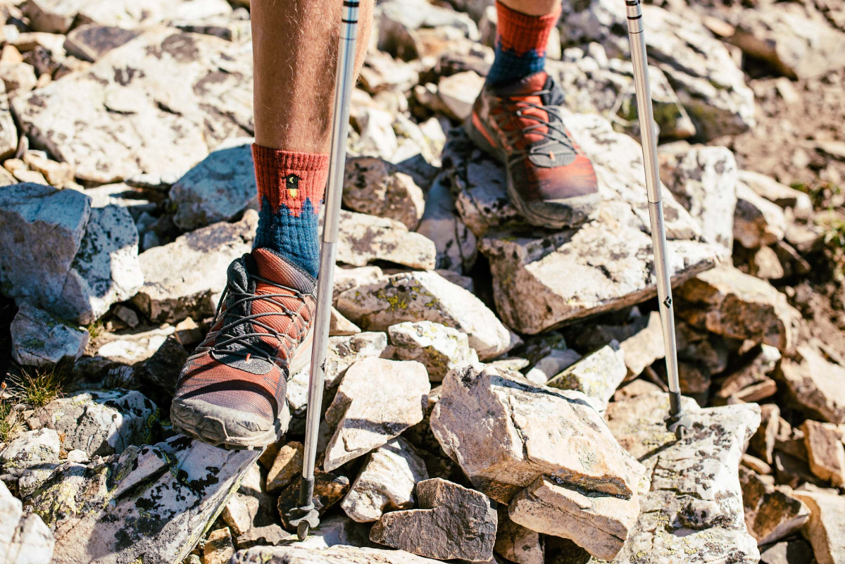 A color photo by Ryan Dearth of man's shoes and hiking poles as he stands on a rocky surface.