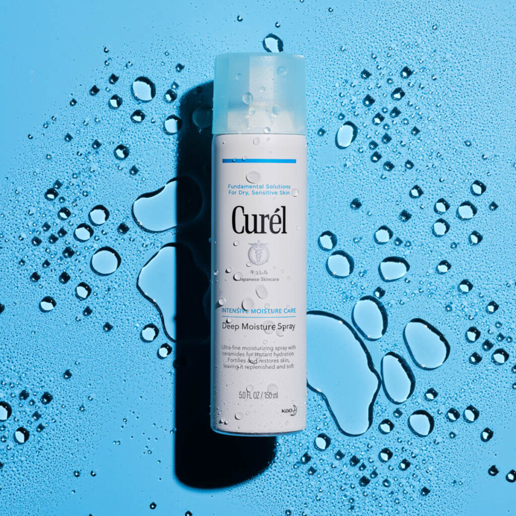 Ryan Kurtz's picture of a bottle of skincare from the brand Curél. The white bottle says "Deep Moisture Spray" and is set against a sky blue surface that is dotted with droplets and small puddles of water. There are water droplets on the bottle as well.