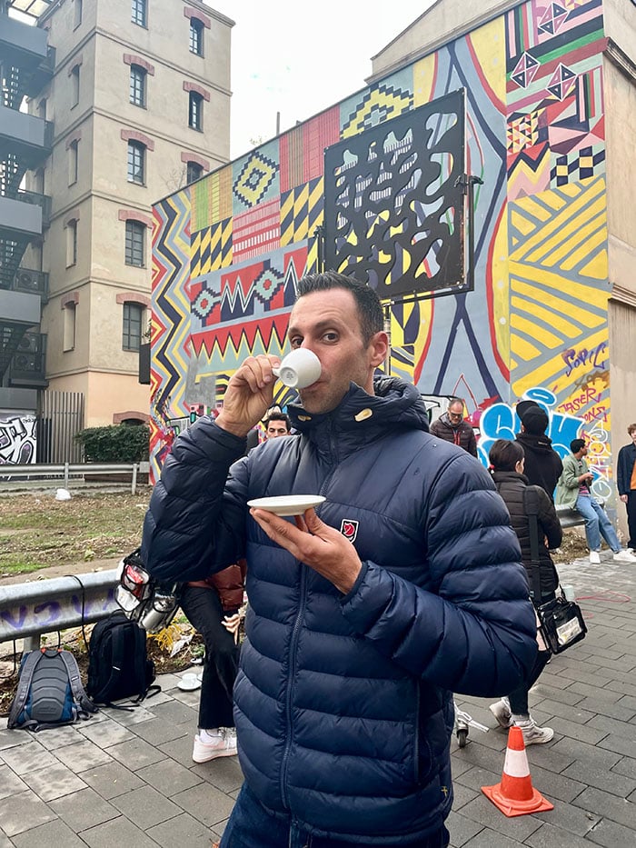 Craig Oppenheimer sipping an espresso while on set. In the background you can see other crew members setting up at a city location with a beautiful graphic mural.