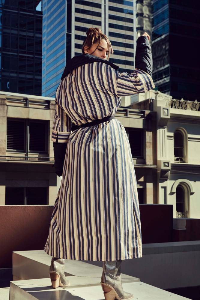 A photo by Steven Popovich of a woman wearing a long striped coat, looking back over her shoulder at the camera on the rooftop in a city center.