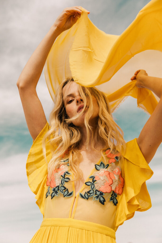 A photo taken by Taryn Kent of a blonde female model wearing a bright yellow dress with pink floral appliqués on the bodice. She waves a translucent yellow scarf over her head with both hands.