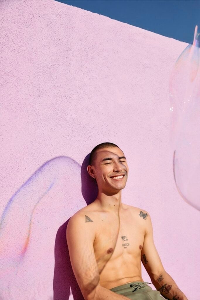 A young man sitting against a pink wall and smiling, photographed by Taryn Kent.
